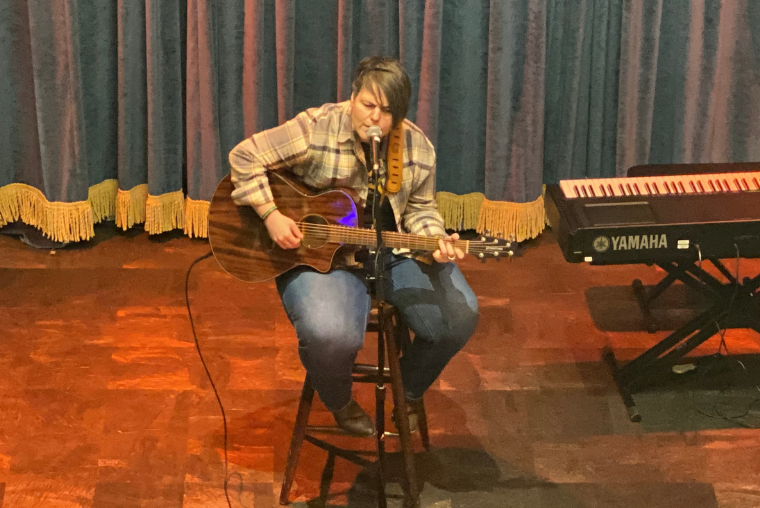 Sundi Jo performing at the Memphis Rock N' Soul Museum, seated on a stool and playing an acoustic guitar, with a Yamaha keyboard in the background on stage.